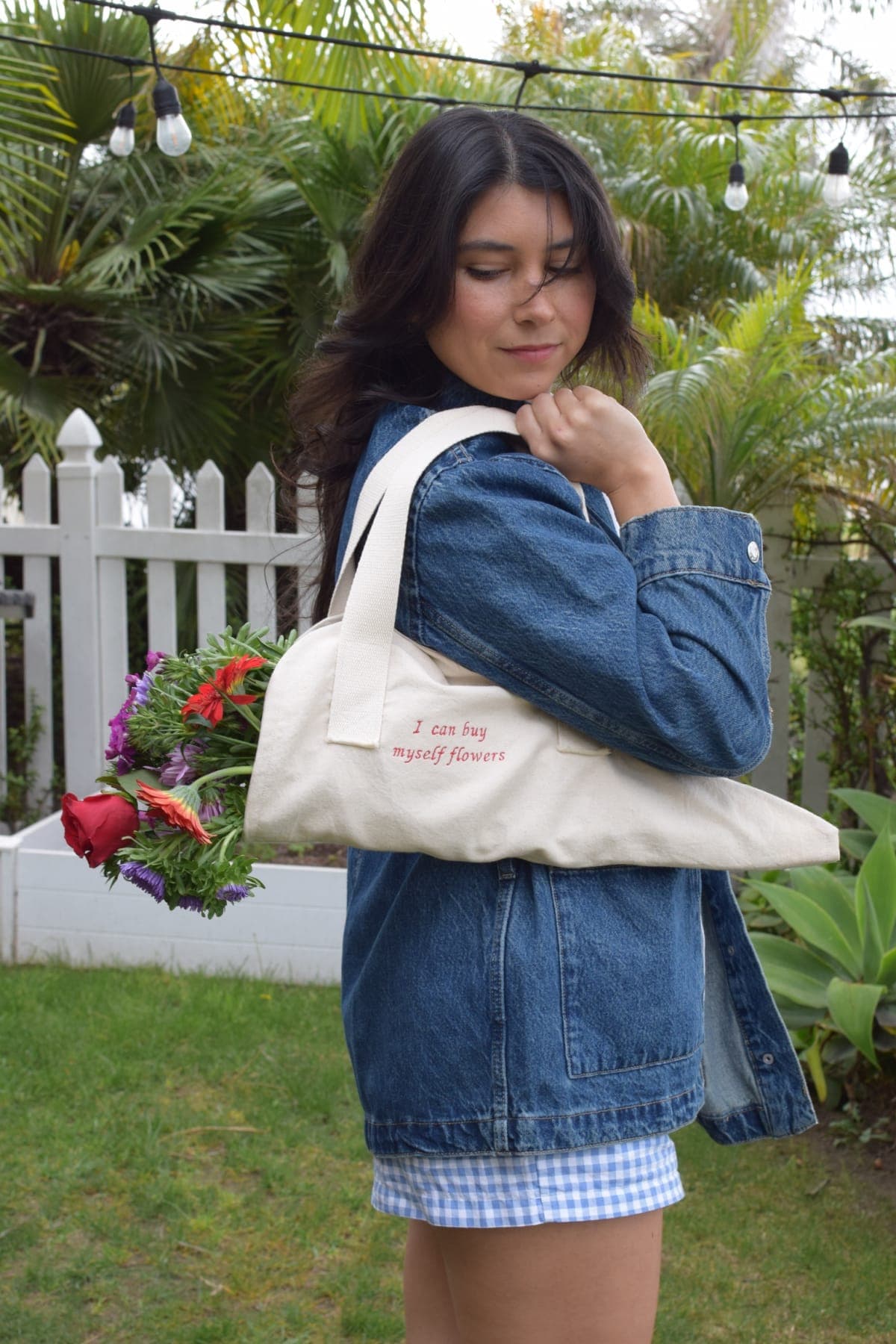 A cream canvas market tote hand-embroidered with "I can buy myself flowers" in coral thread, carried with a fresh bouquet